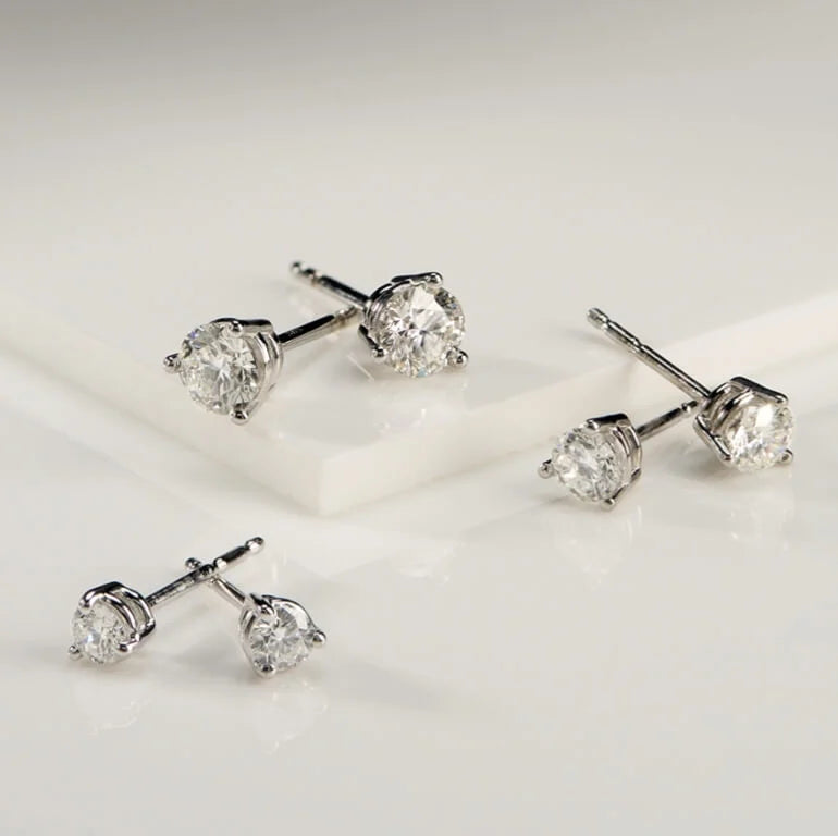 Four round-cut diamond stud earrings in silver settings, arranged on a clean white surface with a white rectangular block.