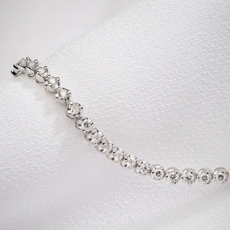 A close-up of a silver tennis bracelet with round-cut clear stones, resting on a textured white fabric surface.