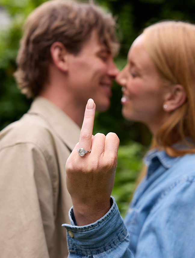 Woman shows off her oval engagement ring as she and her partner lean in to kiss